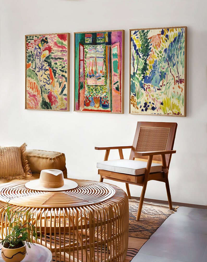 May include: Three framed abstract paintings with colourful brushstrokes depicting a woman, a window with a view of a harbour, and a landscape with trees. The paintings are hanging on a white wall above a wicker coffee table and a wooden chair.