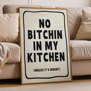 May include: A framed print with the text "NO BITCHIN IN MY KITCHEN (UNLESS IT'S URGENT)" in black on an off-white background. The print has a black border and a wooden frame. The artwork is leaning against a beige sofa.