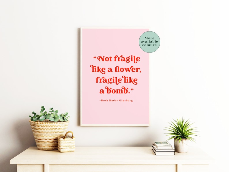 May include: A pink printable quote poster with red text that reads "Not fragile like a flower, fragile like a bomb." - Ruth Bader Ginsburg. The poster is framed and hanging on a white wall.