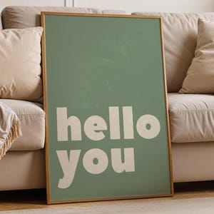 Peut inclure: Une affiche encadrée avec un fond vert sauge et les mots "hello you" en blanc. L'œuvre est présentée contre un canapé beige avec des coussins assortis et une couverture à franges. Le cadre est de couleur marron clair.