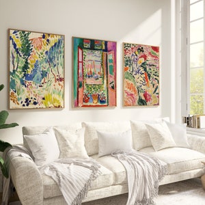 May include: Three framed abstract paintings in a living room setting. The paintings are in a colourful style with a mix of green, blue, pink, and yellow hues. The paintings are hung on a white wall above a white sofa with white throw pillows and a white throw blanket.