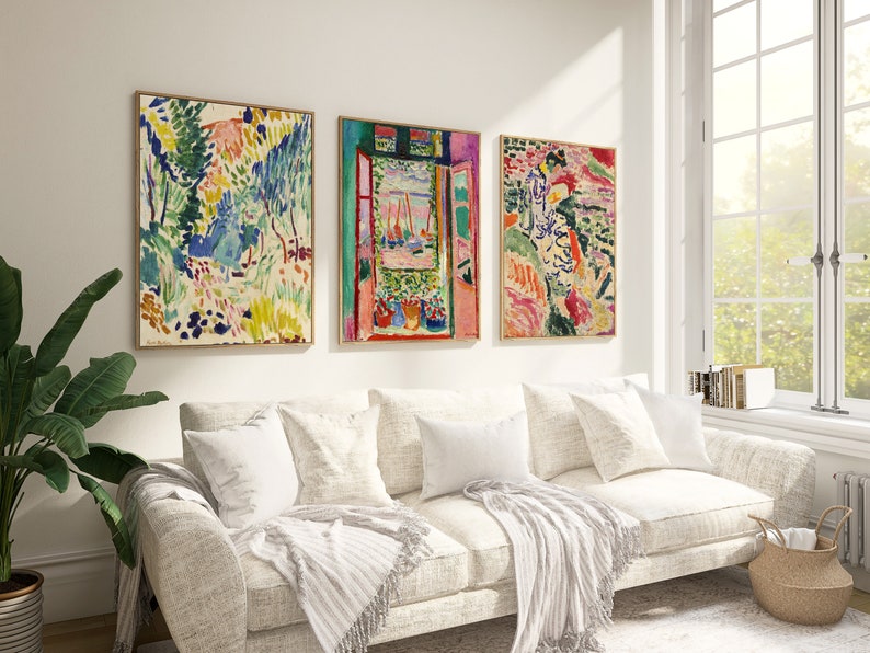 May include: Three framed abstract paintings in a living room setting. The paintings are in a colourful style with a mix of green, blue, pink, and yellow hues. The paintings are hung on a white wall above a white sofa with white throw pillows and a white throw blanket.