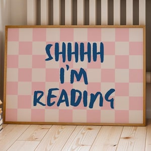 May include: A framed print with a pink and white checkered background. The words "SHHHH I'M READING" are written in blue, bold letters. The print is displayed on a wooden floor.