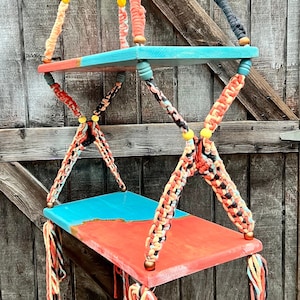 May include: Two turquoise and orange painted wooden shelves hanging from a macrame cord with black, orange, and white tassels. The shelves are suspended from a wooden ceiling.