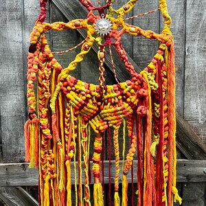 May include: A large macrame wall hanging with a sun design in the center. The macrame is made with red, orange, and yellow yarn. The sun is made with a silver metal charm.