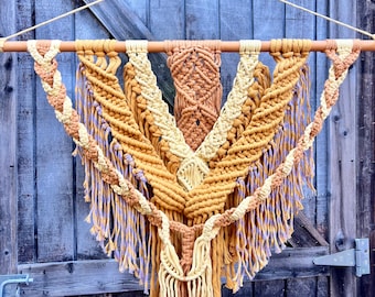 May include: A macrame wall hanging with a geometric design in shades of yellow, cream, and purple. The hanging is made with thick cotton cord and has a fringe at the bottom.