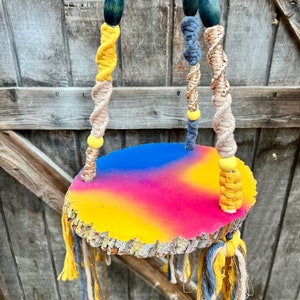 May include: A macrame plant hanger with a wooden, circular shelf painted in a rainbow ombre pattern. The shelf is suspended from the macrame cords with yellow and blue tassels hanging from the bottom.