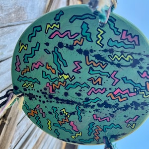 May include: A round, green, painted disc with black paint splatters and a pattern of colorful zigzags. The disc is hanging from a string.
