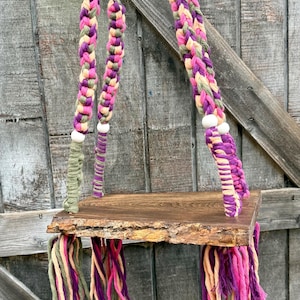 May include: A handmade wooden swing with braided rope supports in shades of purple, pink, green, and beige. The swing's seat is a natural-edged wooden plank, with colorful yarn tassels hanging from the bottom. The swing is suspended against a weathered wooden background.