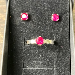 May include: A set of jewelry featuring a ring and a pair of earrings. The ring has a single, large, bright pink gemstone set in a silver band. The earrings are simple studs with a single, bright pink gemstone.