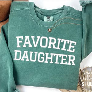 May include: A teal sweatshirt with the words "FAVORITE DAUGHTER" in white block letters. A gold necklace with a small pendant is draped on the sweatshirt. A brown hat is visible in the upper left corner.