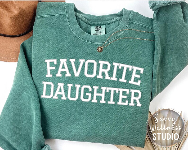 May include: A teal sweatshirt with the words "FAVORITE DAUGHTER" in white block letters. A gold necklace with a small pendant is draped on the sweatshirt. A brown hat is visible in the upper left corner.