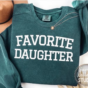 May include: A teal green sweatshirt with the words "FAVORITE DAUGHTER" in white block letters. A gold necklace with a pendant is draped on the sweatshirt. A brown hat is visible in the background.