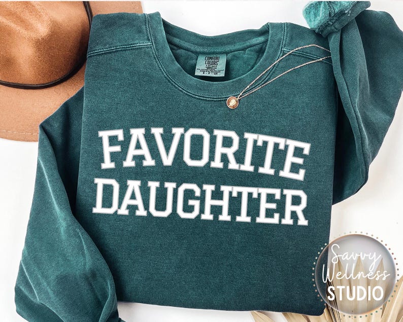 May include: A teal green sweatshirt with the words "FAVORITE DAUGHTER" in white block letters. A gold necklace with a pendant is draped on the sweatshirt. A brown hat is visible in the background.