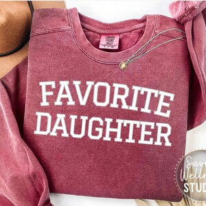 May include: A maroon sweatshirt with the text "FAVORITE DAUGHTER" in white. A gold pendant necklace is draped on the garment. A brown hat is visible in the upper left corner. The Savvy Wellness Studio logo is in the lower right corner.