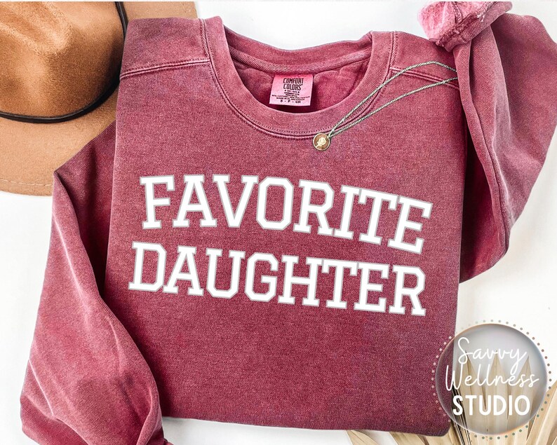 May include: A maroon sweatshirt with the text "FAVORITE DAUGHTER" in white. A gold pendant necklace is draped on the garment. A brown hat is visible in the upper left corner. The Savvy Wellness Studio logo is in the lower right corner.