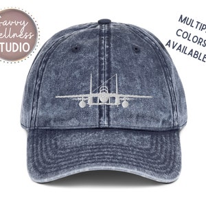 May include: A blue denim baseball cap with a white embroidered silhouette of a fighter jet. The text "MULTIPLE COLORS AVAILABLE!" is printed in white on the right side of the cap.