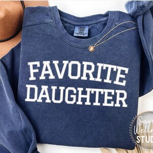 May include: A navy blue sweatshirt with the text "FAVORITE DAUGHTER" in white. A gold necklace with a pendant is draped across the front. A brown hat is visible in the background.