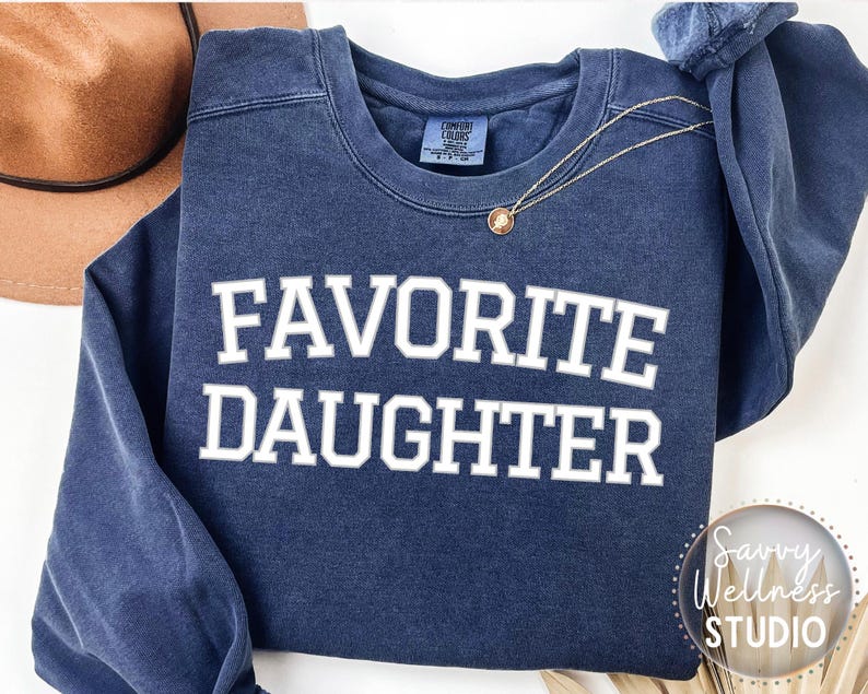 May include: A navy blue sweatshirt with the text "FAVORITE DAUGHTER" in white. A gold necklace with a pendant is draped across the front. A brown hat is visible in the background.