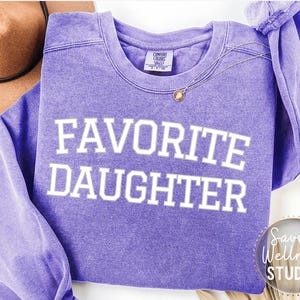 May include: A lilac crew neck sweatshirt with the text "FAVORITE DAUGHTER" in white. A gold necklace with a pendant is draped across the front. A brown hat is visible in the background. The sweatshirt has a vintage, washed appearance.
