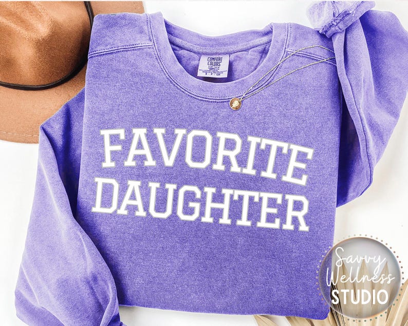 May include: A lilac crew neck sweatshirt with the text "FAVORITE DAUGHTER" in white. A gold necklace with a pendant is draped across the front. A brown hat is visible in the background. The sweatshirt has a vintage, washed appearance.