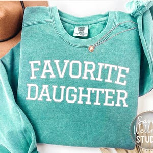 May include: A teal Comfort Colors sweatshirt with the words "FAVOURITE DAUGHTER" in white block letters. A gold necklace with a pendant rests on the shirt. A brown hat is visible in the background. The sweatshirt has a relaxed fit.