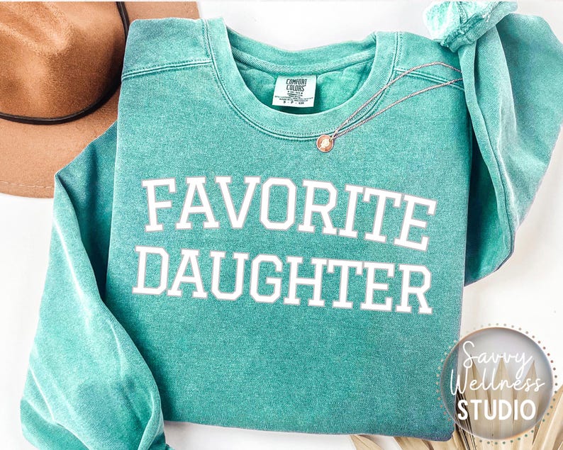 May include: A teal Comfort Colors sweatshirt with the words "FAVOURITE DAUGHTER" in white block letters. A gold necklace with a pendant rests on the shirt. A brown hat is visible in the background. The sweatshirt has a relaxed fit.