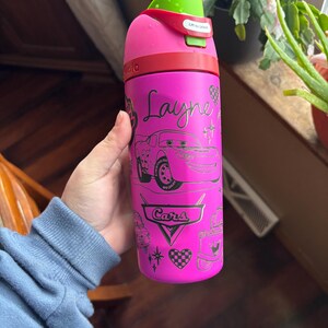 May include: A pink water bottle featuring a Cars theme with the name "Layne" in cursive. The bottle has a green lid and a red band. The design includes illustrations of Lightning McQueen and other Cars-themed graphics.