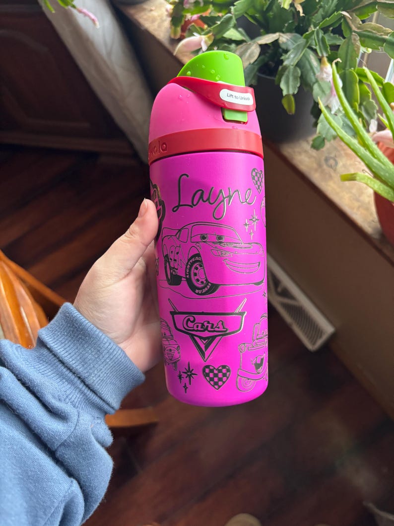 May include: A pink water bottle featuring a Cars theme with the name "Layne" in cursive. The bottle has a green lid and a red band. The design includes illustrations of Lightning McQueen and other Cars-themed graphics.
