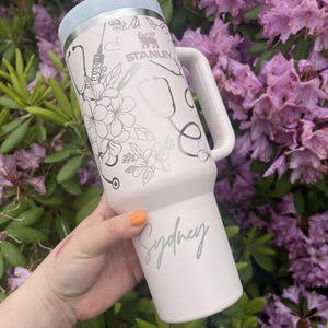 May include: A light pink Stanley tumbler with a light blue lid and handle. The tumbler is decorated with floral and medical-themed graphics, including stethoscopes and syringes. The name "Sydney" is written on the side.