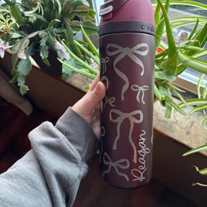 May include: A maroon water bottle with a dark maroon lid and a silver bow pattern. The name "Reagan" is written in a script font on the side. The bottle has a flip-top lid and the brand name "owala" is visible.