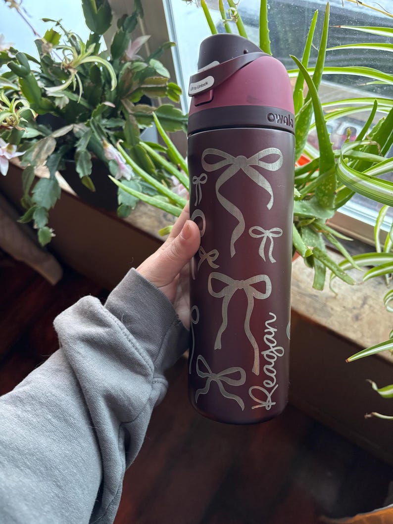 May include: A maroon water bottle with a dark maroon lid and a silver bow pattern. The name "Reagan" is written in a script font on the side. The bottle has a flip-top lid and the brand name "owala" is visible.
