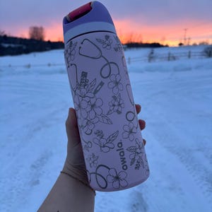 May include: A light pink Owala water bottle with a purple lid, decorated with a floral and medical theme. The design includes stethoscopes, thermometers, and flowers. The background shows a snowy landscape and a sunset.