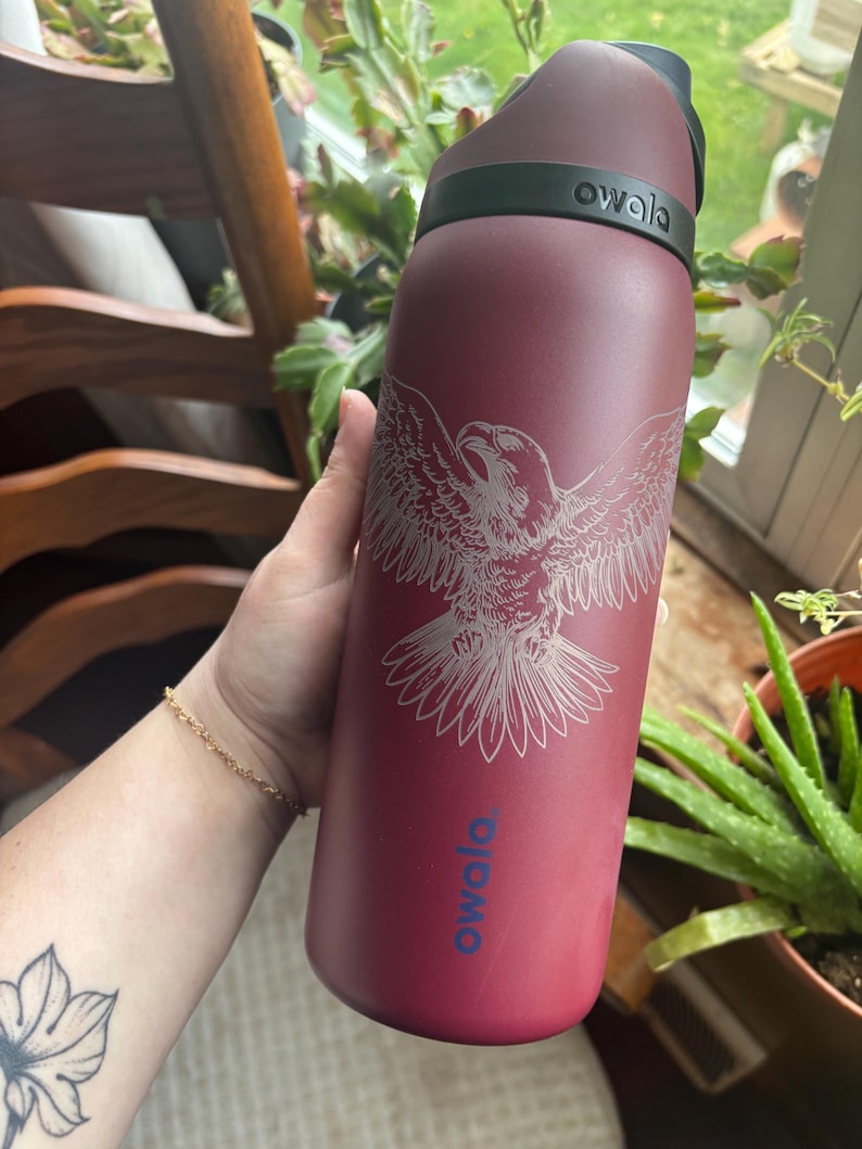 May include: A maroon Owala water bottle with a silver eagle design. The bottle has a black handle and a black lid. The Owala logo is printed in blue. The bottle is held in front of a window with plants.