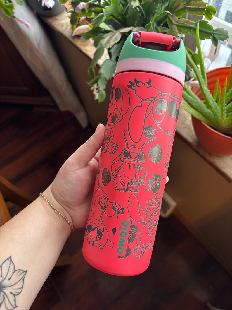 May include: A red Owala water bottle featuring a design of the character Stitch from Lilo & Stitch. The bottle has a green and pink lid. The bottle is covered in black line art of Stitch and tropical flowers.