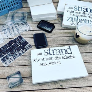 May include: A white wooden sign with black text that reads "am strand zieht man die schuhe aus, wie man das zuhause eben auch tut". The sign is sitting on a wooden table with other crafting supplies, including ink pads, alphabet stamps, and a glass of milk.