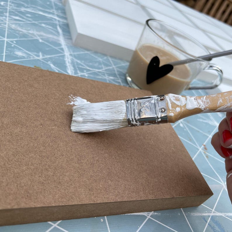 May include: A white paint brush with white paint on it, being used to paint a brown wooden surface.