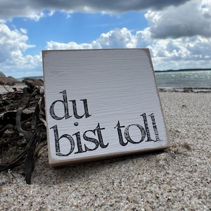 May include: A white wooden sign with black lettering that reads "du bist toll" on a sandy beach.