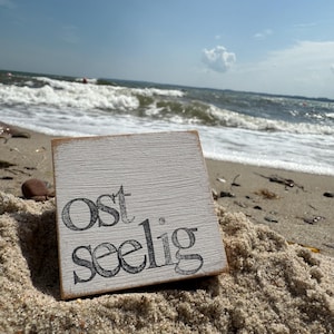 Könnte beinhalten: Ein quadratisches, verwittertes Holzschild mit dem Wort "Ost Seelig" in schwarzer Schrift, das auf einem Sandstrand liegt. Das Schild ist weiß gestrichen und hat eine rustikale, Vintage-Ästhetik. Der Ozean und der Himmel sind im Hintergrund zu sehen.