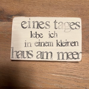 May include: A white wooden sign with black lettering that reads "eines tages lebe ich in einem kleinen haus am meer".