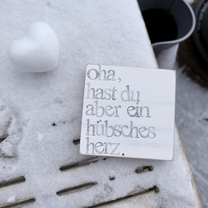May include: A white, heart-shaped object sits next to a small, white wooden sign with black text. The sign reads "Oha, hast du aber ein hübsches herz." The scene is set on a snow-covered wooden surface.