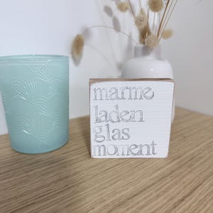 May include: A square white wooden block sign with distressed edges featuring the silver text marme, laden, glas, moment stacked vertically. Beside it sits a light blue glass candle holder with a textured fan pattern on its exterior surface.