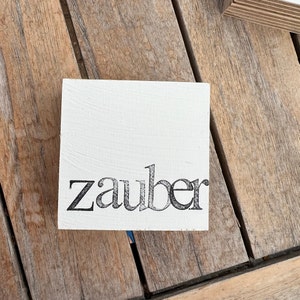 Könnte beinhalten: Ein quadratischer, weißer Holzblock mit dem Wort "zauber" in schwarzer, stilisierter Schrift. Der Block liegt auf einer Holzoberfläche.