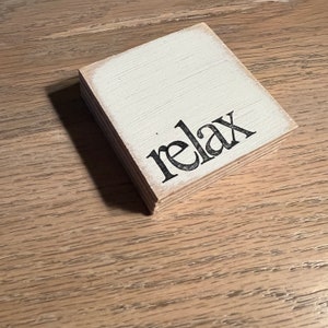 May include: A small, square wooden block with a distressed white painted surface. The word "relax" is printed in black, stylized lettering. The block is made of layered wood and sits on a light brown wooden surface.