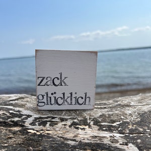 Könnte beinhalten: Ein weißes Holzschild mit schwarzem Text, auf dem "zack glücklich" steht, auf einer Holzoberfläche.