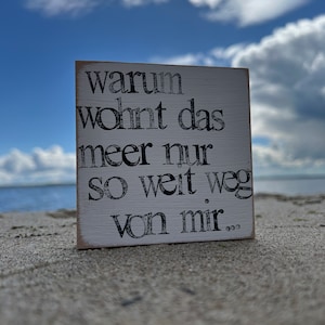Könnte beinhalten: Ein kleines, weißes Holzschild mit schwarzem Text, das auf Sand ruht, mit blauem Himmel und Wasser im Hintergrund. Auf dem Schild steht: "warum wohnt das meer nur so weit weg von mir..."