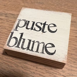 May include: A white wooden sign with black lettering that reads "puste blume".