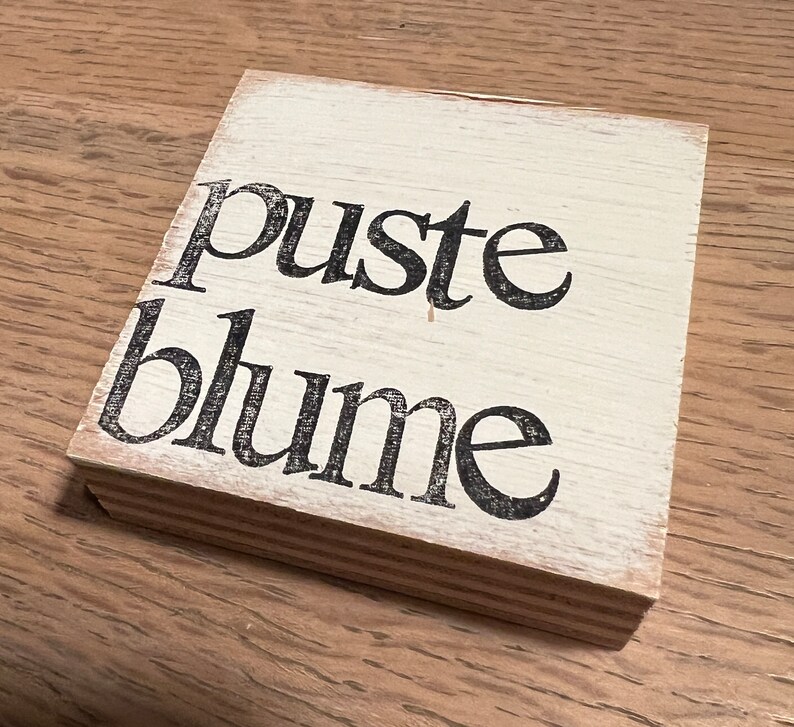 May include: A white wooden sign with black lettering that reads "puste blume".