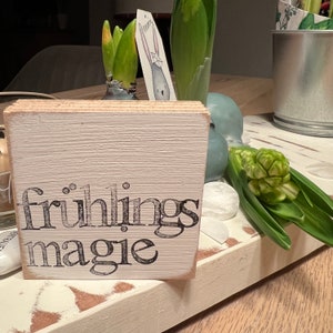May include: White wooden sign with black lettering that reads "fr&uuml;hlings magie". The sign is sitting on a whitewashed wooden surface with green hyacinth flowers and a white ceramic object.