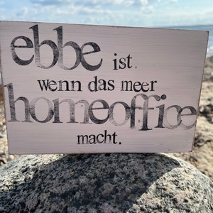 Könnte beinhalten: Ein weißes Holzschild mit schwarzem Text, der "ebbe ist, wenn das meer homeoffice macht." lautet. Das Schild liegt auf einem grauen Stein.